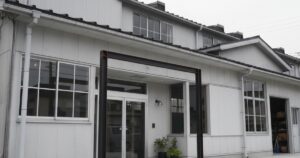 白い外壁の建物の正面入口。大きな窓とガラス扉が並び、シンプルで工場併設オフィスのような外観。